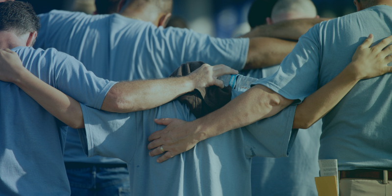 A group of people in blue shirts with arms around each other's shoulders, viewed from behind